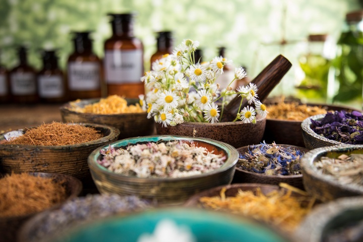 Assorted dried herbs and flowers in wooden bowls, with essential oil bottles in the background, on a table.