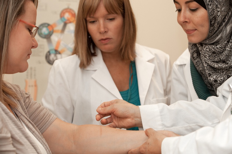 A medical professional inserts a needle into a patient's arm while two other professionals observe.