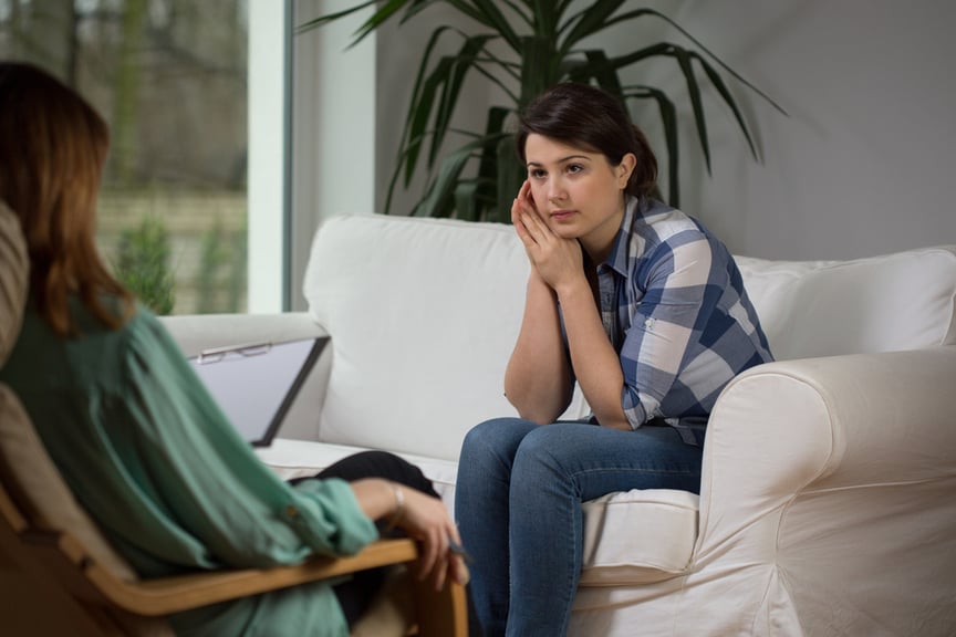 A woman in a plaid shirt sits on a white couch, engaged in conversation with another person holding a clipboard in a bright room with large windows and plants.